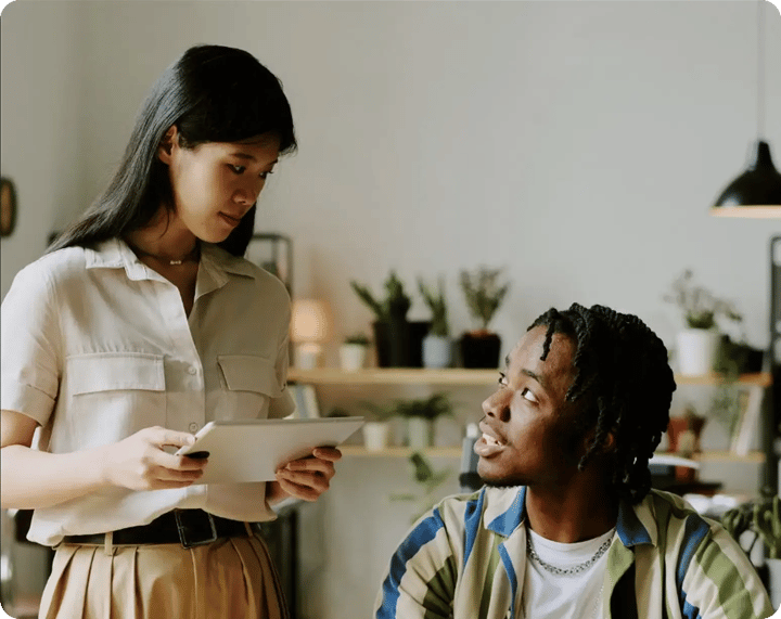 A young woman holding a tablet points out information to a young Black man sitting at a desk in a brightly lit room as they discuss content together.