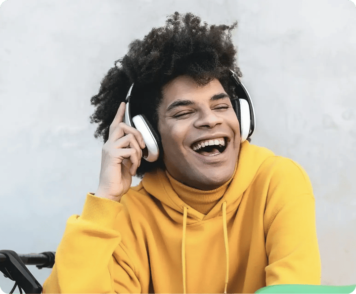 Close-up photo of a cheerful young man wearing a mustard yellow hoodie and over-ear headphones, laughing outdoors.