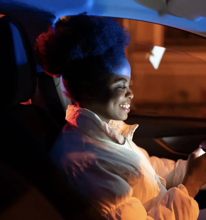 Profile photo of a young Black woman with styled hair smiling happily from the front seat of a car at night under soft, colorful ambient lighting.